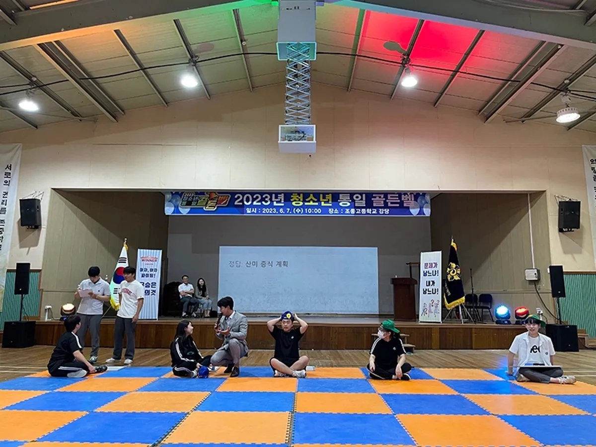 강당 바닥에 체크무늬 매트를 깔고 무대 앞에서 진행하는 실내 행사 장면