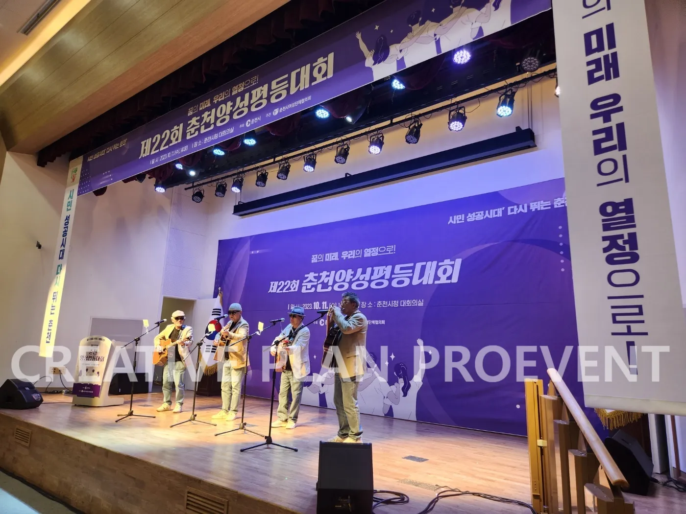 보라색 대회 현수막을 건 강당 무대에서 기타 공연팀이 노래하고 위쪽에 조명이 달린 모습